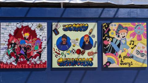A selection of artwork displayed on a hoarding outside a construction site. The art on the left shows a band and says 'Youth Arts Jersey'. The second painting shows two smiling characters and says 'Jersey Grainville'. The third is mostly yellow and pink and hows a boy playing with a ball. Words over the top read 'A place 4 every1'.
