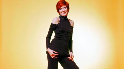 A young Cilla Black poses in front of a yellow background. She wears a black jumpsuit with cut out shoulders and a high neck. She has short red hair.