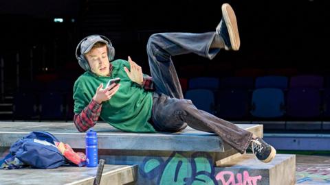 Dan Rainford in Top G's Like Me. The actor, wearing a green baggy polo and blue jeans is laying on his side in a skatepark and scrolling on his phone as he listens to content through headphones.