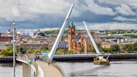 A view of Londonderry with the Peace Bridge over the Foyle river in the foreground and the Guildhall and other building in the background.
