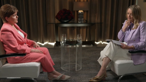 Two people sit across from one another in what looks like a calm, staged interview setting. One wears a pink suit with a white top, sitting upright with hands resting together. The other wears a light‑purple blazer and white trousers, holding a notepad and pen as if guiding the conversation. Between them is a small glass table with two glasses of water. Behind them, a console table holds a lamp and a bowl of red flowers, framed by long curtains 