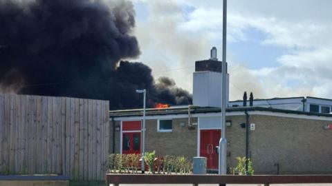 A school building on fire. Thick black smoke billows out of the roof of the building.