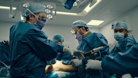 Doctors and nurses wearing blue scrubs and facemasks and gloves. They are carrying out surgery on a patient.