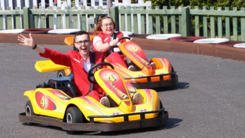 A young man beams widely at the camera has he drives a yellow go-kart around a track. He is waving with one arm towards the camera. He is wearing a red jacket and trousers. To the right, a woman with ginger hair and glasses, also dressed in red, drives a similar car. 