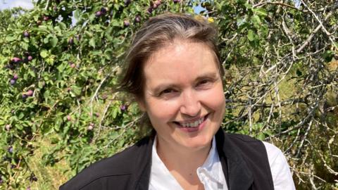 A woman wearing a white shirt and a black gilet is standing next to a hedgerow which includes damson trees. She has brown hair and is smiling for the photograph.