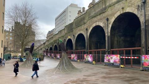 Pavemented area in the foreground with two artworks in the shape of electrical cables with railway arches in the background. 