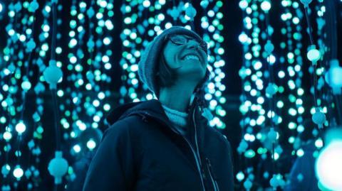 The picture shows a person standing in the middle of an immersive light installation. The scene is filled with vertical strands of glowing blue and white lights, creating a sparkling, almost underwater-like atmosphere. The lights appear to be suspended from above in dense clusters, giving the impression of walking through a tunnel or forest of illuminated orbs. The person is dressed in a winter jacket and a knitted hat.