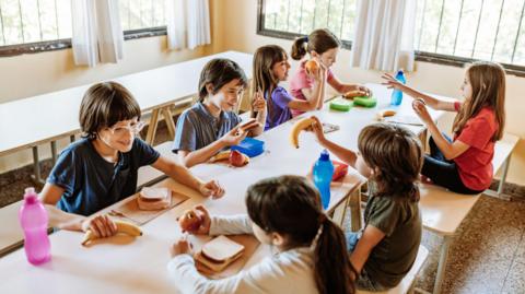 Children sat around a table at a breakfast club. There are seven children, all eating packed lunches. 