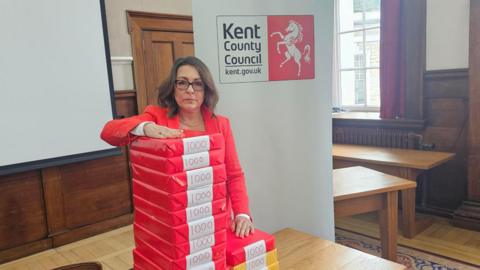 Kent County Council leader Linden Kemkaran, wearing a red jacket, leans on a large pile of packets of A4 paper piled on a table at County Hall in Maidstone, each one labelled with the number 1,000.