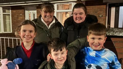 three children and two women smiling for the camera. One of the children is wearing a football themed tracksuit, one is wearing a thick rain coat, and the other is wearing a school uniform.