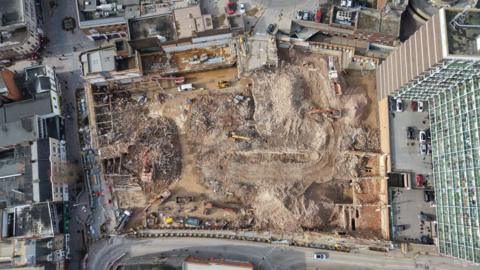 An aerial shot from directly above the site shows buildings fully demolished with some debris and mounds of dirt. There are also construction vehicles. 