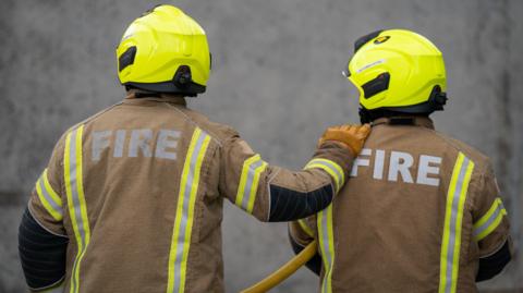 The backs of two firefighters wearing brown fire jackets and wearing yellow helmets. One is holding a yellow hose under their left arm while the other has their hand on the other's shoulder.