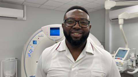 Bempah photographed looking into the camera and smiling. He is wearing a white collared shirt with a dark red trim around the collar, and dark-framed glasses. He has short, dark curly hair and a short beard. He is pictured inside an MRI scanning room, with the machine - a large, white, doughnut-shaped structure - behind him. The machine has an array of blue lights on it, and a screen at the apex. The room has dark grey walls and a white ceiling. 