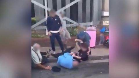 Two older men dressed in blue and green lying on the floor while three men in darker colours stand over them