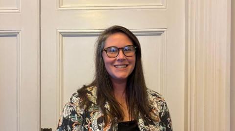 Charlotte Traynor, a woman with long dark hair, she wears large round glasses and has a bright smile, she wears a patterned white and black jacket.