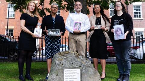 Four women and one man stand alongside a large rock showing a memorial plaque. They are all holding photos of relatives killed in road crashes.