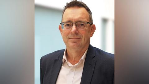 Paul Simpson wearing glasses, a shirt and a blazer and looking at the camera.