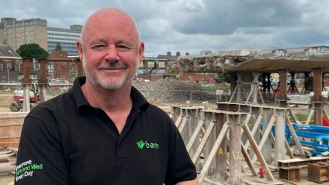 Jim Symth, a middle-aged man with a shaved head and a white stubble-beard, wearing a BAM construction contactor polo shirt. In the background is the jagged edge of the thick, concrete flyover, which has been demolished up to this point. Steel posts stick out in all directions, like the end of a frayed rope, which run through the concrete to reinforce it. Beneath the bridge is earthy ground, surrounded by a chainlink fence.