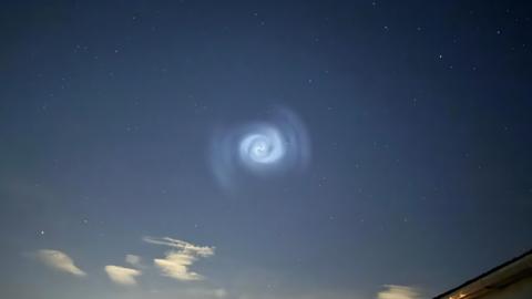 Pale blue glowing spiral shape in the night sky. It has cloud-like markings around it. The middle is particularly bright.