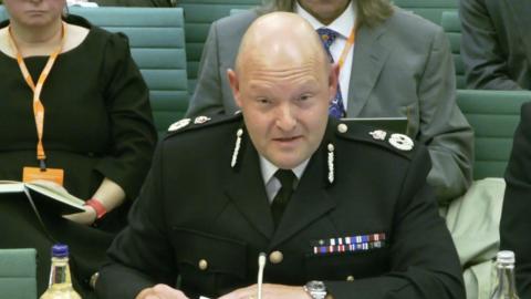 Former Chief Constable Craig Guildford sits at a desk while wearing a black police uniform with a medal ribbon on it. Several people are sitting on green chairs behind him.