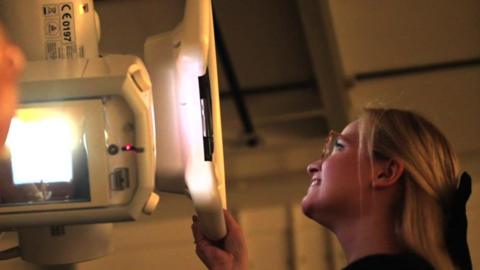 Jo Shuttlewood-Limb operates a scanning machine. She is smiling into the machine as it works and she views what it is doing. She has long blonde hair with a black bow in it at the back of her head.