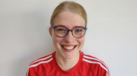 Jess Lowe, a 25-year-old white woman with strawberry blonde hair which is tied up. She is wearing a red Adidas T-shirt and glasses with a brown and blue frame. 