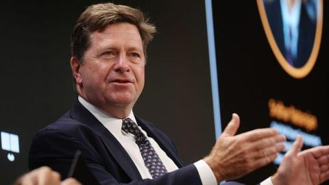 Jay Clayton wearing a suit and raising his hands as he speaks at an event