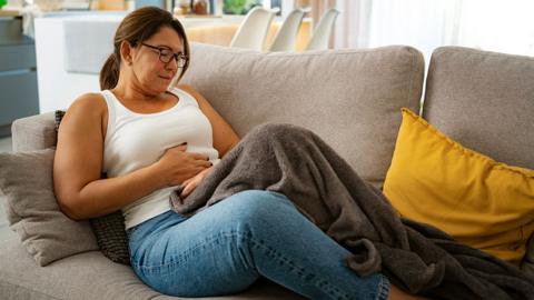 A woman sits on the sofa at home with a blanket on top of her and yellow cushion to her side, clutching her stomach.