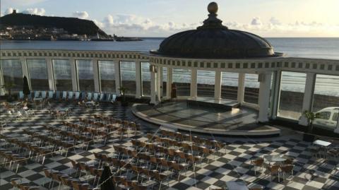 Seating laid out on a black and white checked floor space inside Scarborough Spa.. The headland, sea and beach are in the background.