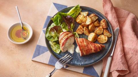 A blue plate filled with a cooked chicken breast wrapped in bacon, crispy potatoes and salad leaves