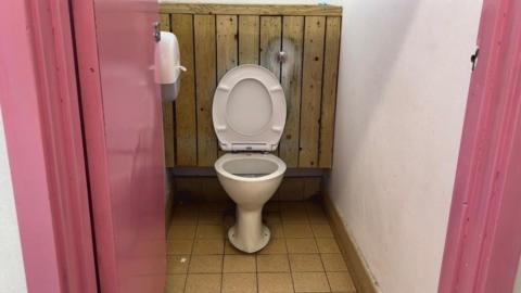 Toilet cubicle and the door and frame is pink. The floor is a bit dirty