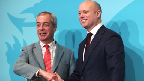 Two men in suits in front of Blue Reform-branded background, shaking hands