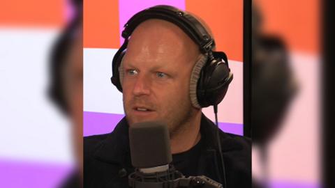 Man with stubble, wearing headphones talks into a mic in a radio studio.