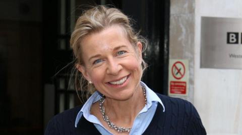 Katie Hopkins stood outside a BBC building wearing a navy blue jumper with a light blue shirt underneath. She is stood smiling at the camera with her blonde hair tied in a bun