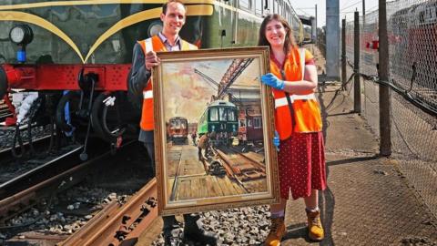 Two rail workers stand on tracks holding painting