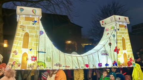 A paper lantern model of Clifton Suspension Bridge, illuminated from the inside, carried by a few people on their shoulders