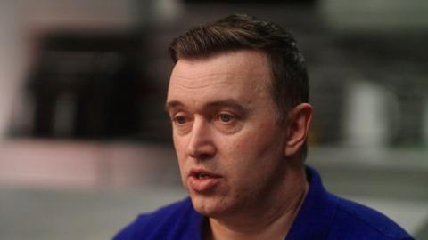 Séamus Flannigan wearing a royal blue top, pictured from the shoulders up. He has brown hair. The background of the image is blurred. 