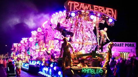 A lit up carnival cart at Bridgwater carnival in 2023. The lights are pruple, red and blue. There are dressed up people standing on it and a crowd around the cart.