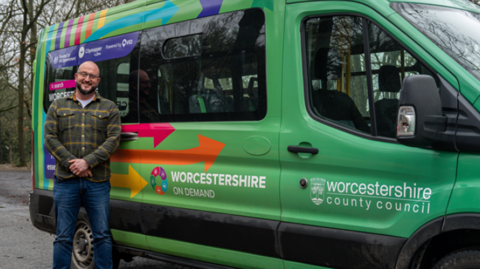 Councillor Karl Perks , a man in blue jeans and a chequred shirt is standing next to a green bus with the words, Worcestershire on demand across the side