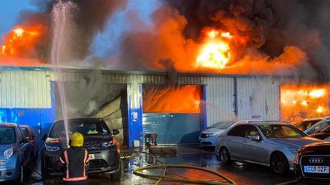 A firefighter can be seen directing a hose onto the roof of a metal warehouse building. Flames are shooting from the door and the roof. A number of cars are parked outside the building