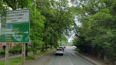 A road, with cars lining up at traffic lights, heading away from the camera. To the left are houses, trees and a large green road sign, while to the left there are trees and a thick green hedgerow