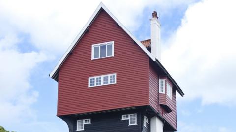 Close-up of The House in the Clouds, a water tower disguised as a house. The camera is at ground level, looking up and a red boarded house with white chimney and a black tower below, set against a blue sky with white clouds.