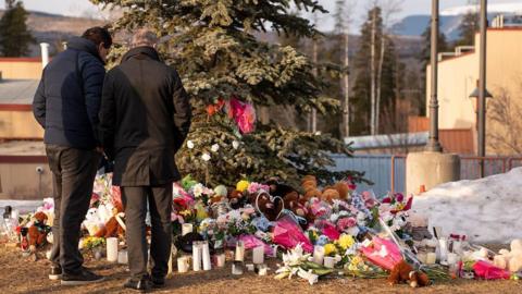 Two men standing outside, backs to camera looking down at a large collection of flowers and presents left for the victims of the school shooting, placed underneath a large tree.