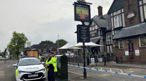Police officer stands by a police car in front of a police cordon around the Fairway Inn in New Moston following the death of a boy.