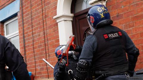 A police officer in blue helmet and riot gear batons down a brown door at the front of a redbrick house while anoth officer in riot gear watches on