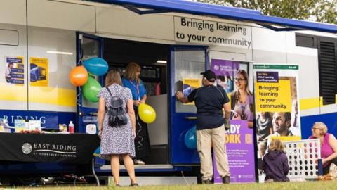 A white bus is stationary with two blue doors opening up on its side. It has the words 'bringing learning to your community' in font on the side. The back of a woman can be seen stood outside it, with two other people sat on the floor next to a large connect four game. Balloons are tied to the doors with a table in front which says 'East Riding of Yorkshire Council' on it. A woman in a blue top and lanyard is stood at the entrance to the bus.