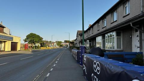 A road with houses along one side and a used car business on the other. There are soft crash barriers outside the house. A red phone box and tree can be seen in the distance. The sky is clear.