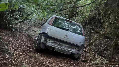 A silver Fiat Punto car left damaged and abandoned in a woodland area. The car is missing its rear bumper and number plate and is amongst trees and branches. 