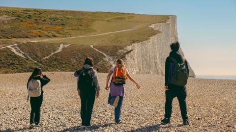 Rear view of a group of hikers walking across a beach toward white cliffs on a sunny day