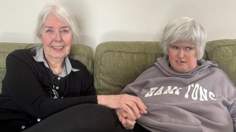 Judith and Monica are sitting on a green couch. Judith on the left has grey hair and is smiling at the camera, she has glasses on a chain round her neck. She is wearing a black cardigan with a black and white striped shirt underneath, she's holding her daughter's hand. She has grey hair and is wearing a dusty lilac hoodie with the word "Hamptons" written in white across it.